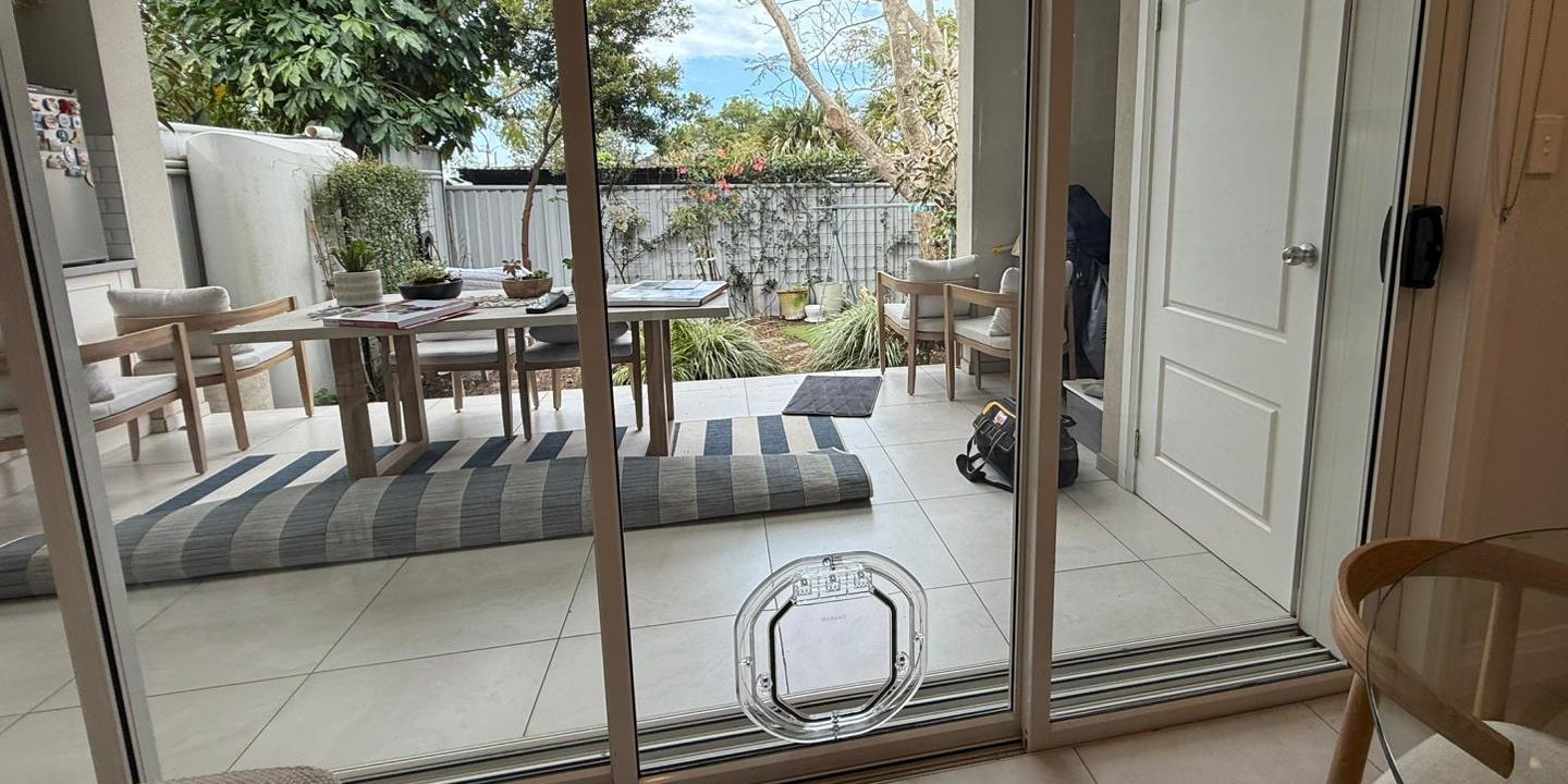 Clear glass-fitting pet door fitted low in an aluminium sliding door, looking out to a covered patio and garden